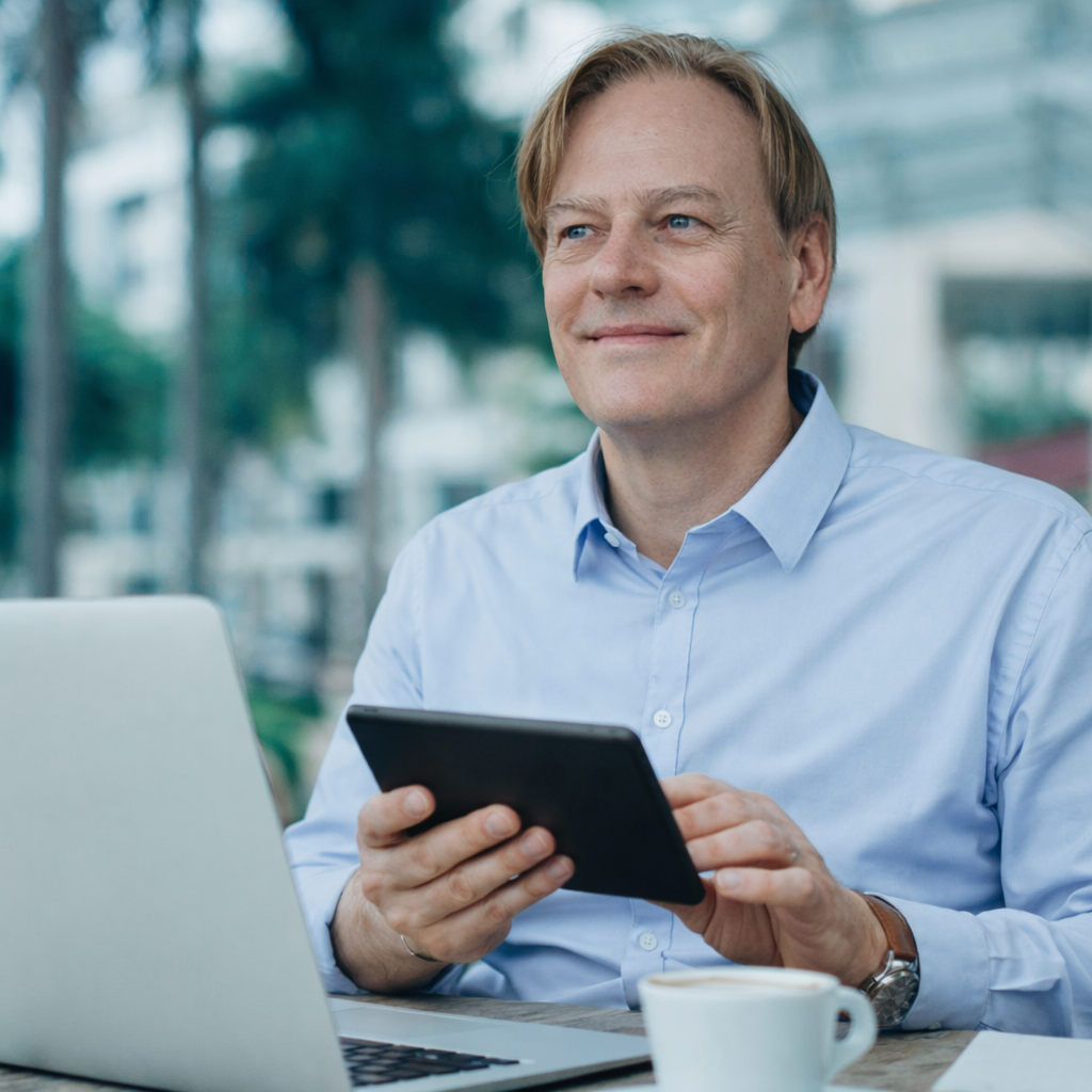 Olivier Dupont, Expert en vidéo LinkedIn et Marque personnelle Homme avec une tablette, souriant, assis à une table à l'extérieur avec un ordinateur portable.